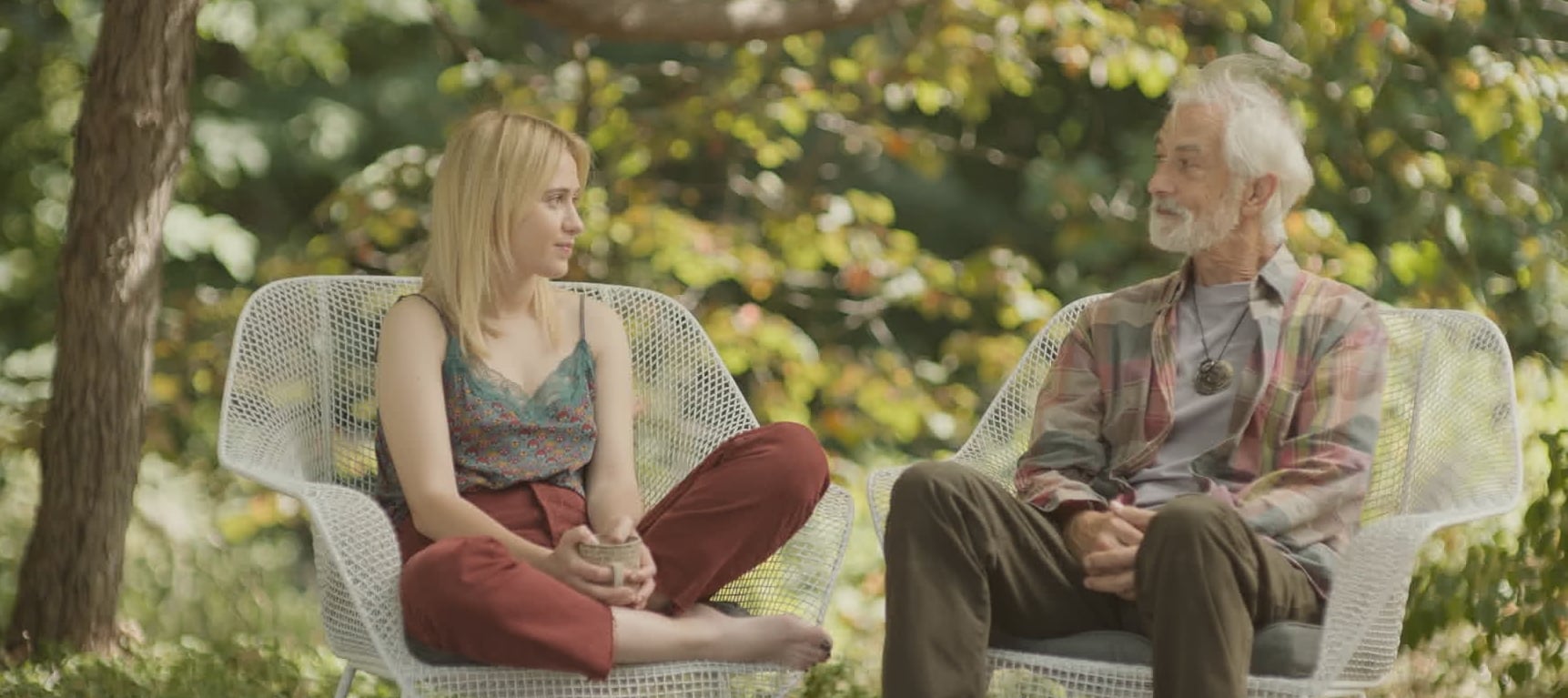 A young woman and an older man sit on white outdoor chairs facing each other, talking in a leafy garden setting.