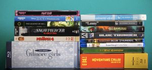 No Streaming Required: Physical Media Spotlight For The Week Of May 12th A stack of assorted DVD and Blu-ray cases, including titles like "Fight Club," "Snowpiercer," "Gilmore Girls," and others, arranged on a turquoise background.