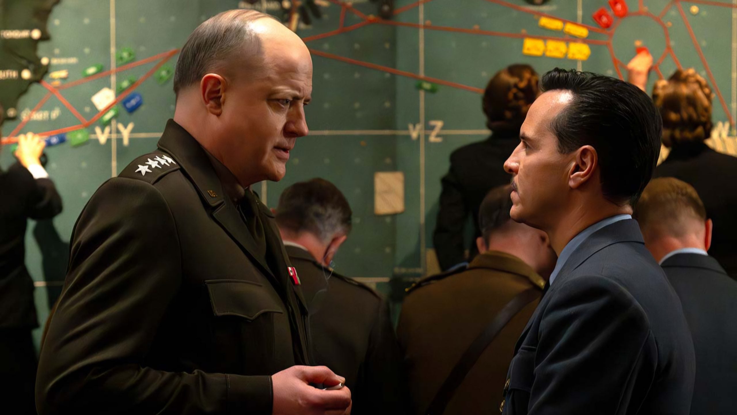 Two men in military uniforms have a serious conversation in front of a large strategic map covered with lines and markers, while other uniformed individuals work in the background.