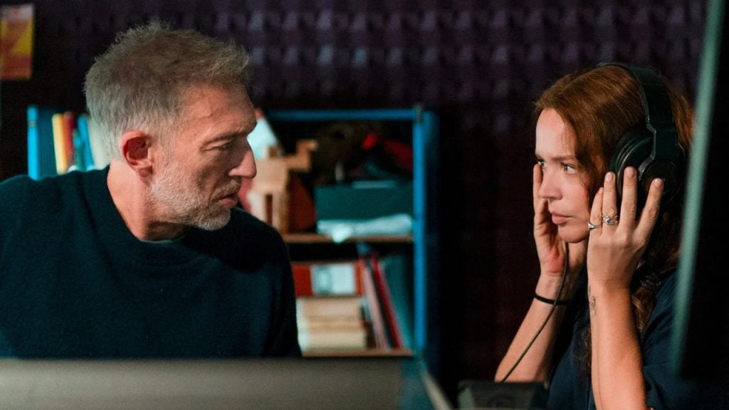 A man and a woman sit indoors; the woman wears headphones and looks focused, while the man looks at her and speaks. Shelves with books are visible in the background.