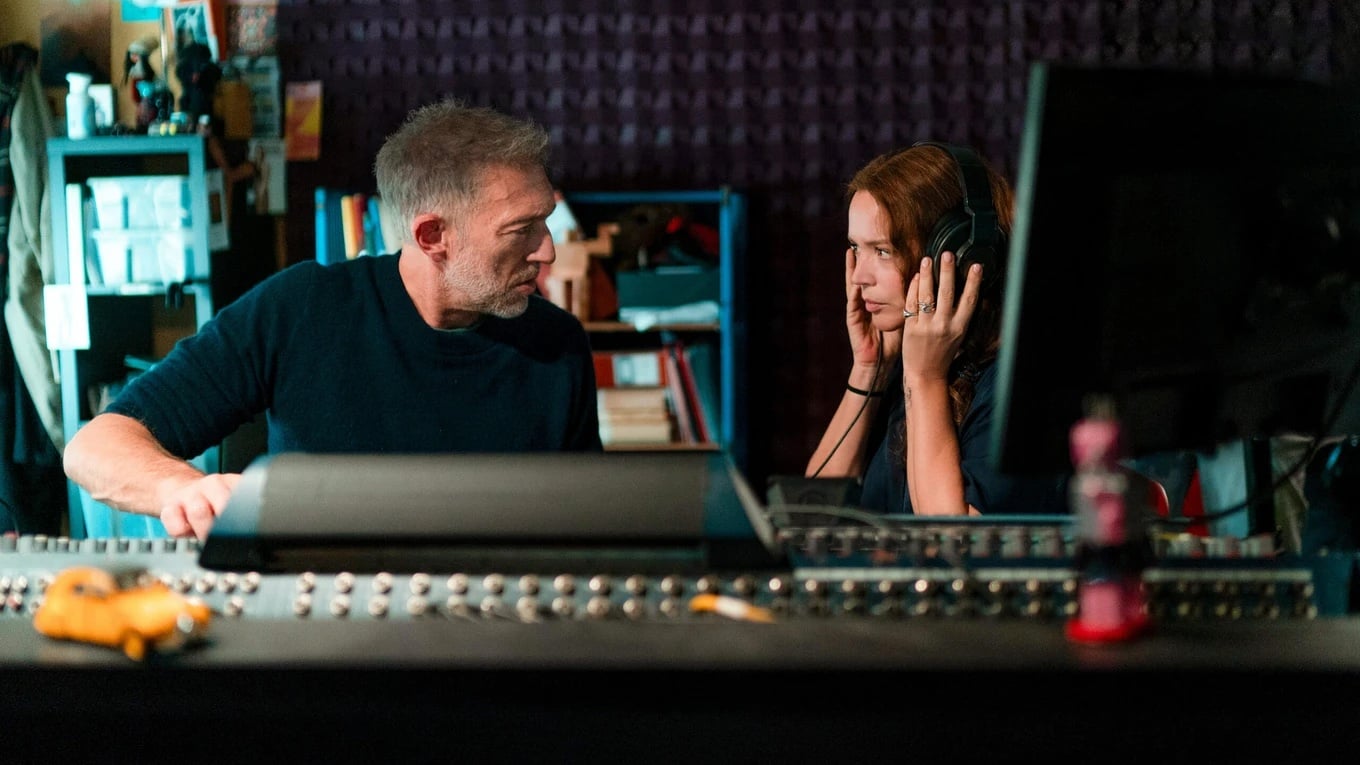 Two people sit in a recording studio; one is wearing headphones while the other looks at them, surrounded by audio equipment and shelves.