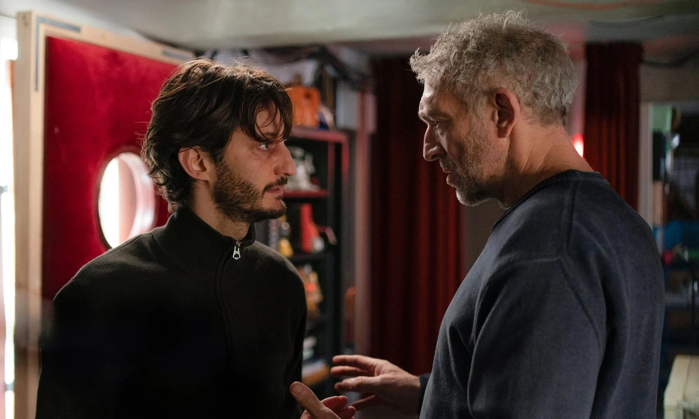 Two men are standing indoors, facing each other and having a serious conversation. One gestures with his hands while the other listens intently.