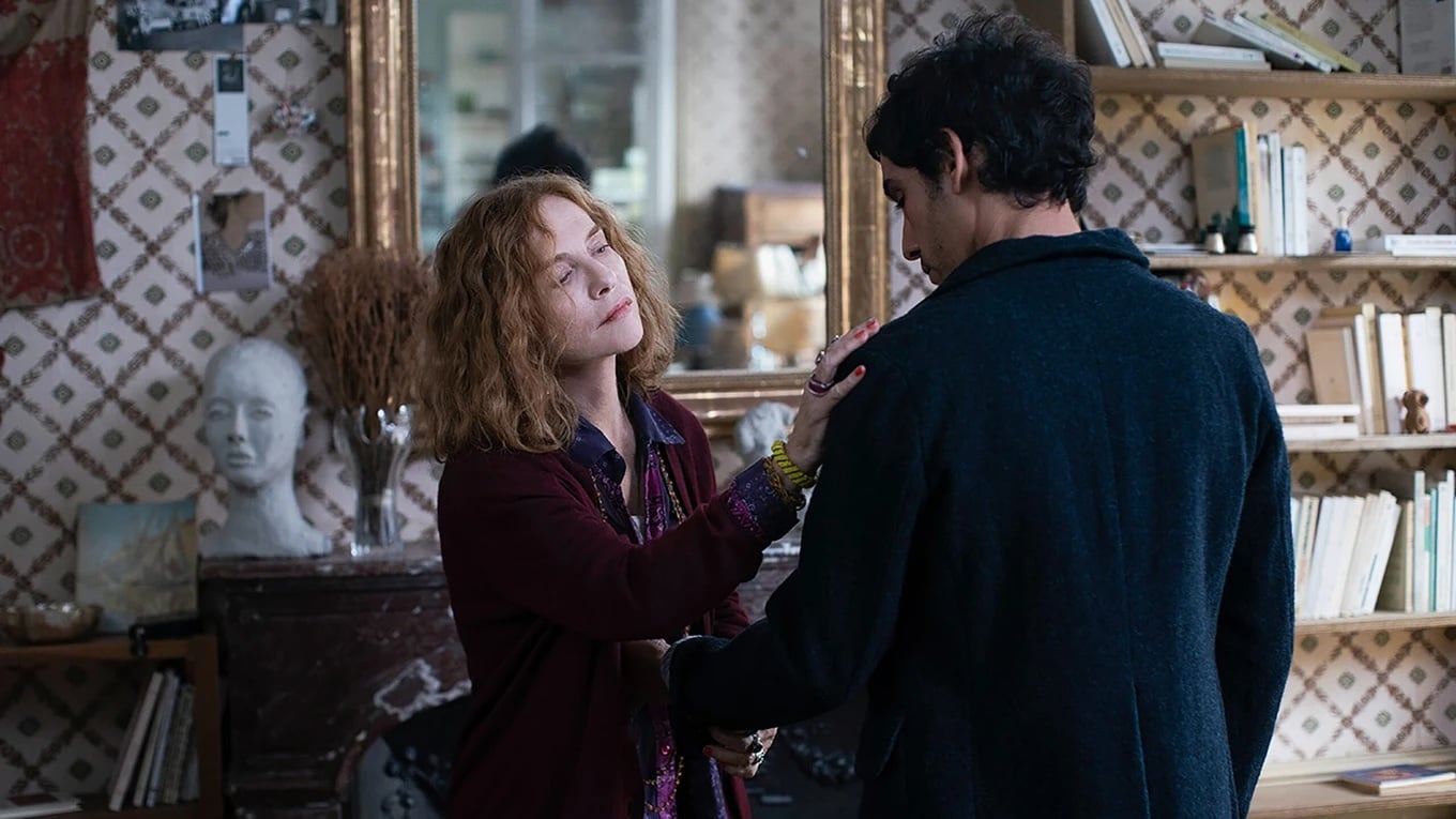 A woman with curly red hair gently places her hand on a man's shoulder in a cozy, book-filled room with patterned wallpaper and a large mirror.