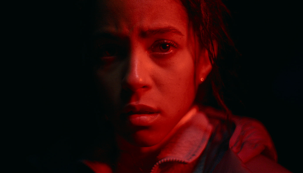 A young person with a serious expression is illuminated by red light against a dark background.