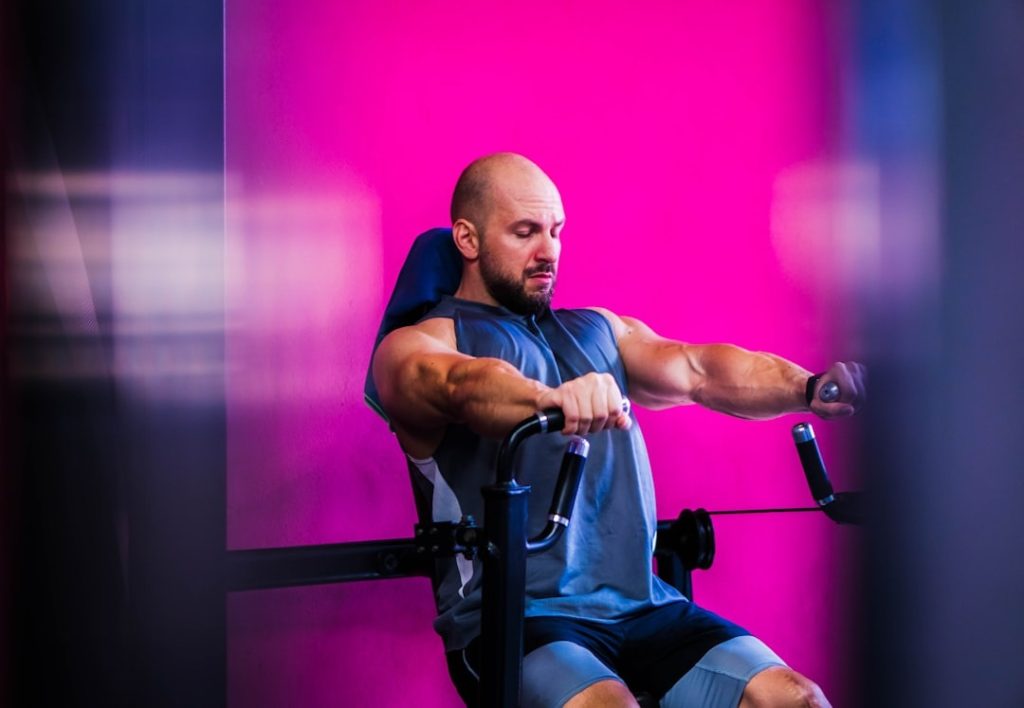 A man wearing a sleeveless shirt uses a rowing exercise machine in a gym, with a bright pink wall in the background.