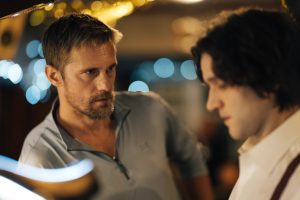 A24 To Release The Alexander Skarsgård & Harry Melling Leather-Clad Romance ‘Pillion’ On Blu-Ray This July A man with short hair and a beard looks intently at another man with longer hair in a dimly lit setting with blurred lights in the background.