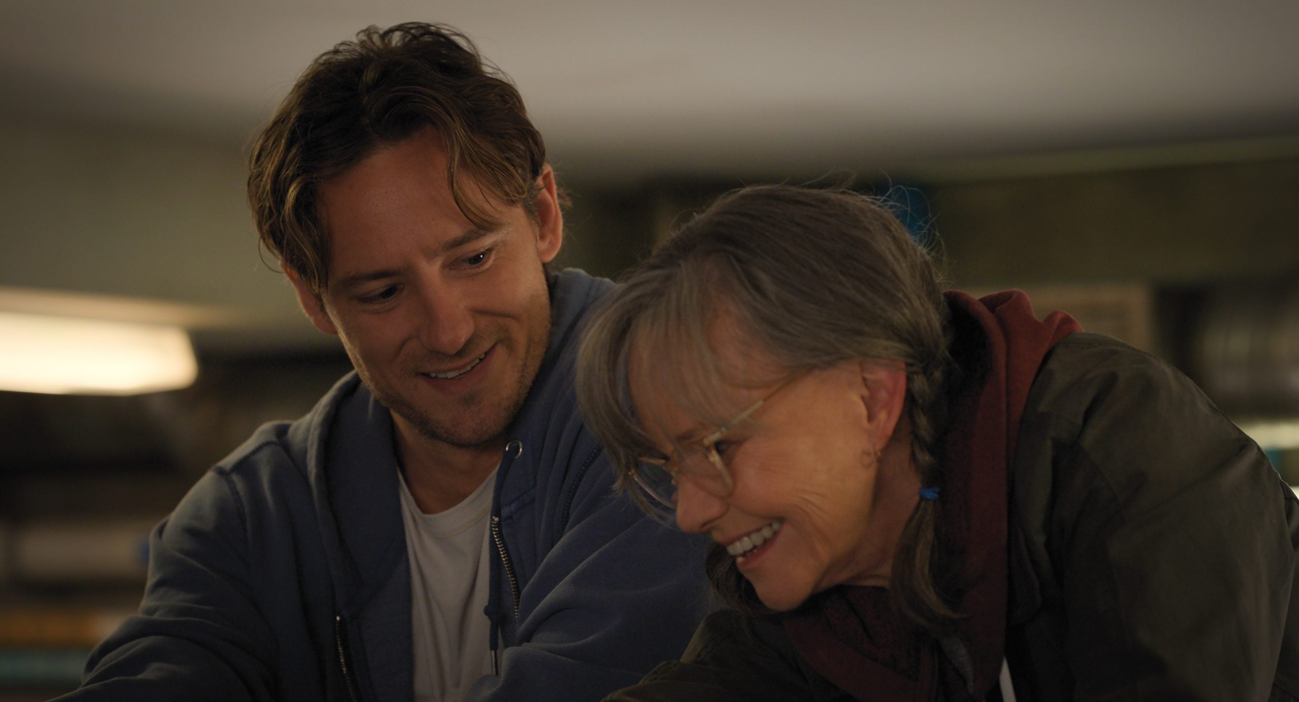 A man and an older woman smile and look down together in a warmly lit indoor setting.