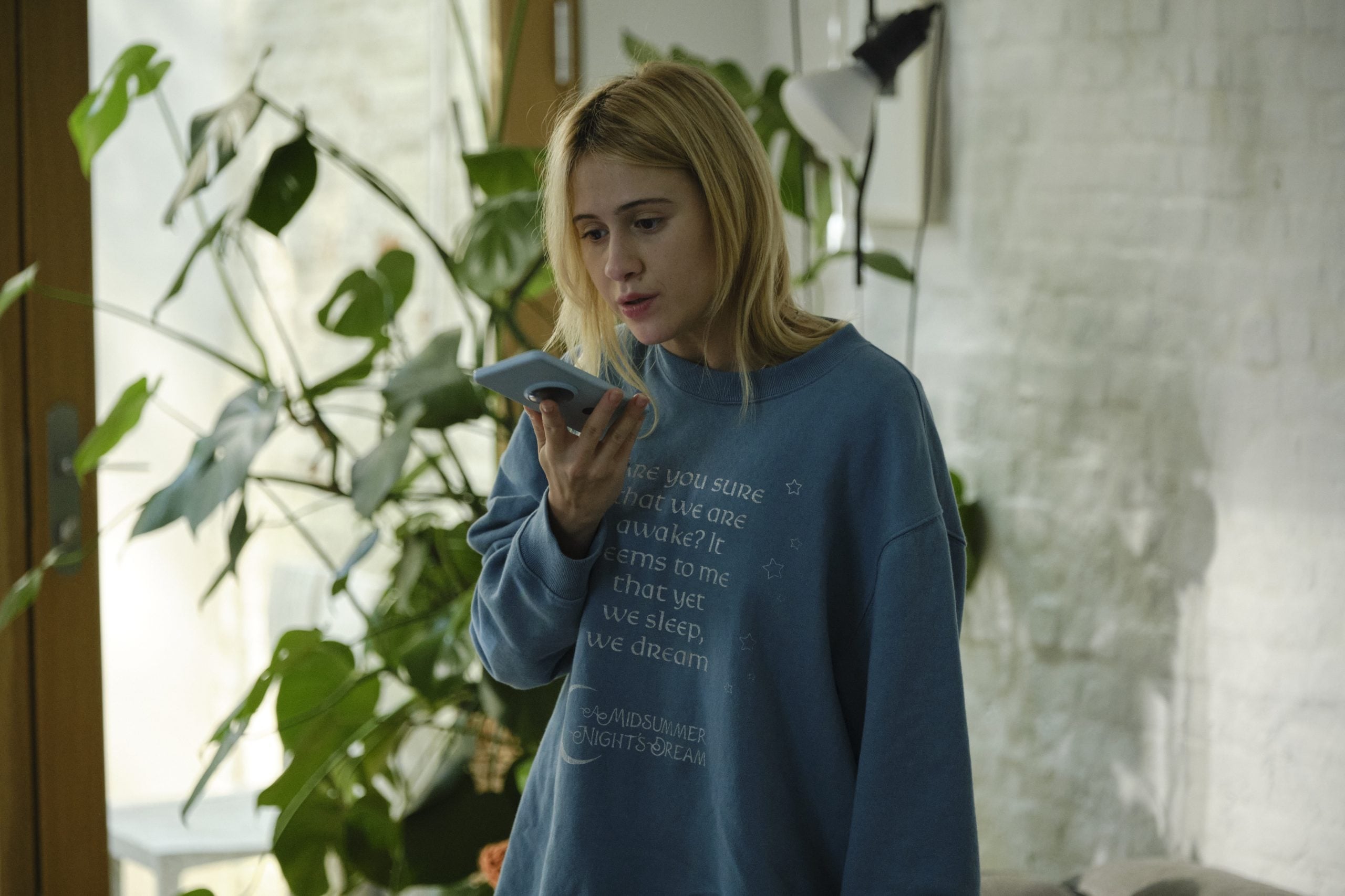 A person in a blue sweatshirt is speaking into a smartphone while standing indoors near large plants and a window.