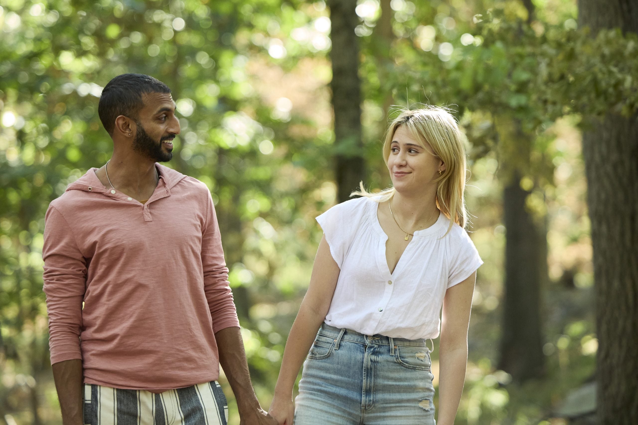 A man and a woman walk hand-in-hand through a wooded area, looking at each other and smiling.