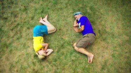 A young girl and an older man lie on grass, positioned to form the shape of the letter "Q" with their bodies.