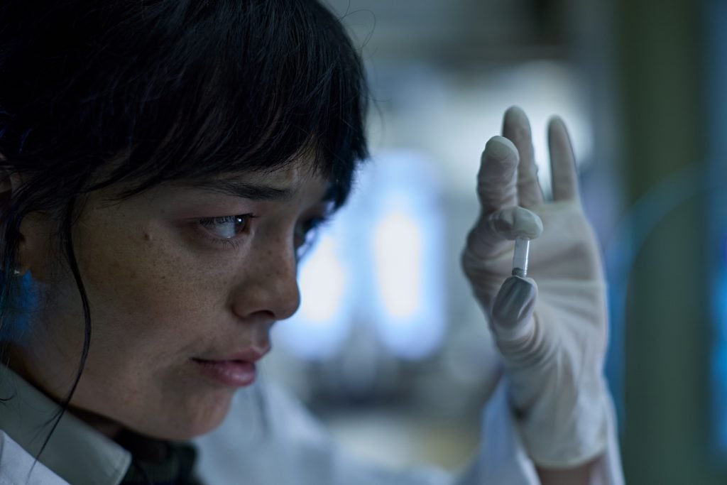 A scientist wearing gloves holds up a small vial, closely examining its contents in a laboratory setting.