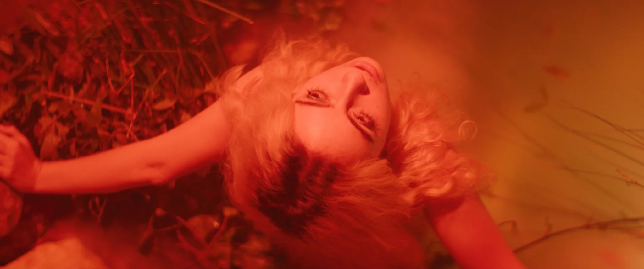 A person with curly blonde hair lies on the ground, looking up toward the camera, surrounded by foliage and bathed in warm orange-red light.