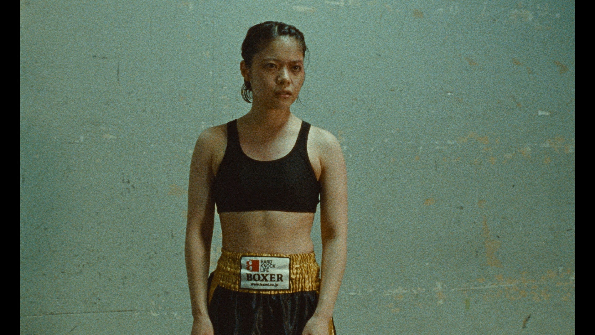 A woman in a black sports bra and boxing shorts stands against a plain, worn wall, looking forward with a serious expression.