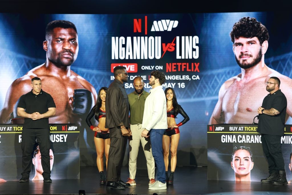 Two fighters face off on stage at a weigh-in event, with promotional screens and staff members standing nearby in the background.