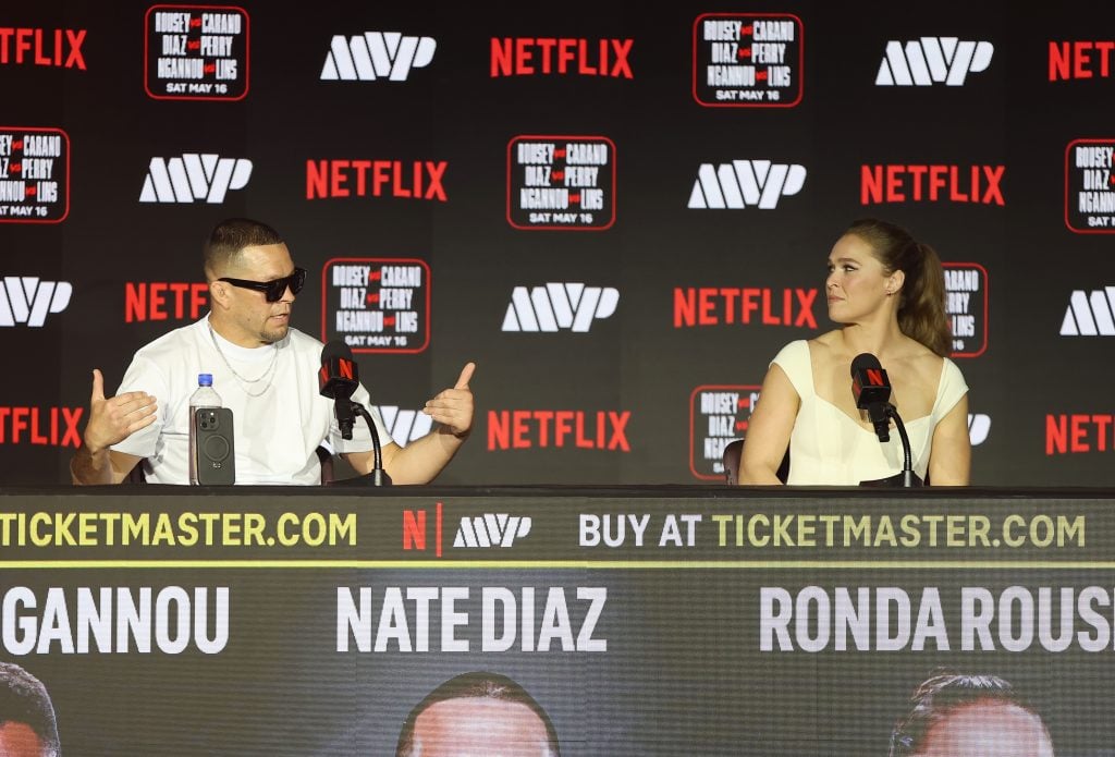 Nate Diaz speaks at a press conference table with Ronda Rousey, surrounded by sponsor logos and event signage.