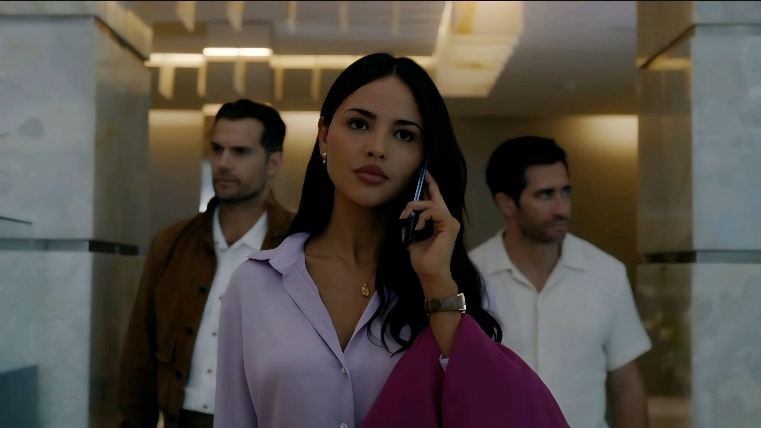 A woman in a lavender blouse talks on her phone while walking in a modern building, with two men following behind her.