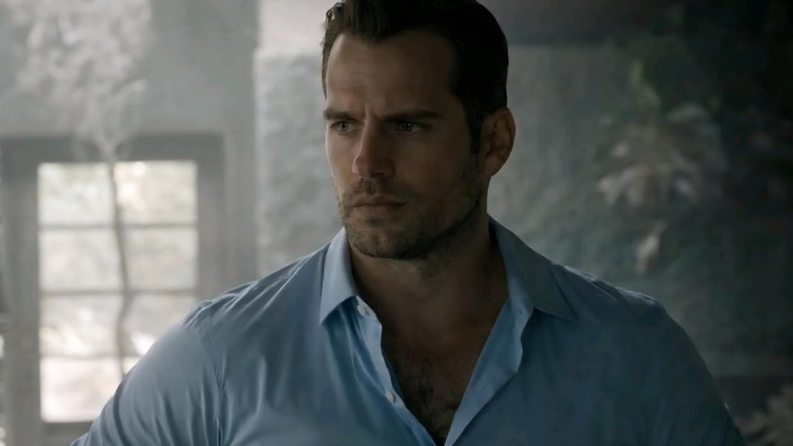 A man with short dark hair and stubble wearing a light blue button-up shirt stands indoors, looking to the side with a serious expression.
