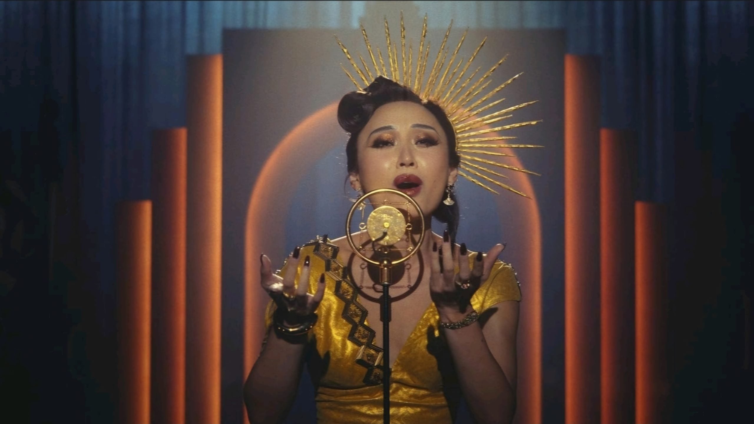 A woman in a gold dress and sunburst headpiece sings into a vintage microphone on stage with art deco-style lighting in the background.