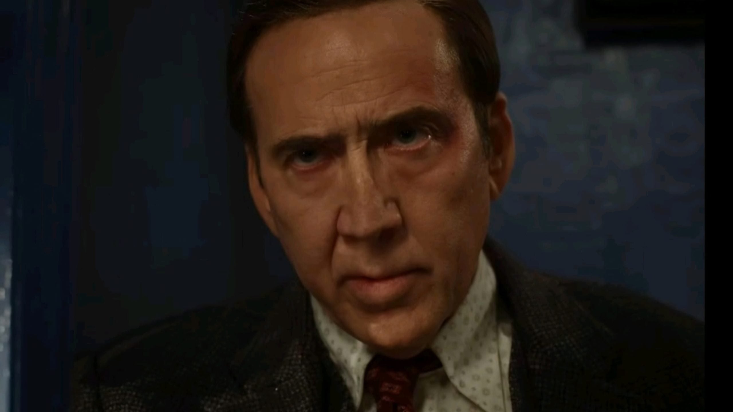 A middle-aged man in a suit and tie stares intently forward with a serious expression against a dark, blurred background.