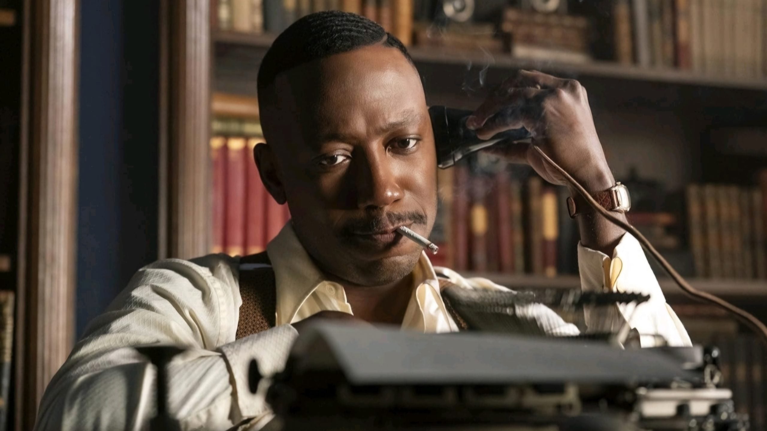 A man in a dress shirt sits at a desk with a typewriter, holding a phone to his ear and a cigarette in his mouth. Bookshelves filled with books are visible in the background.