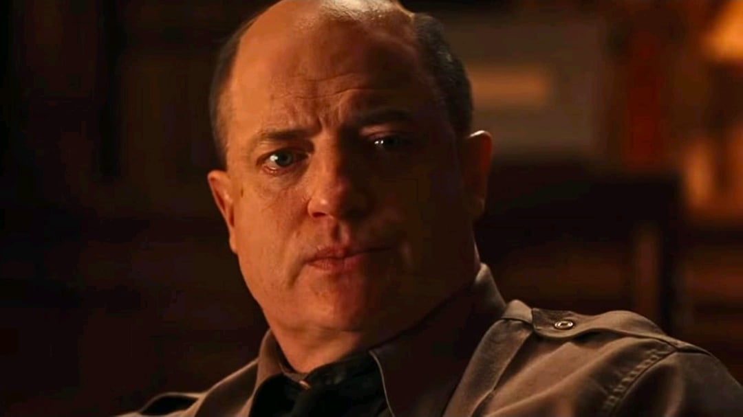 A middle-aged man in a brown uniform shirt sits indoors, looking forward with a serious expression.
