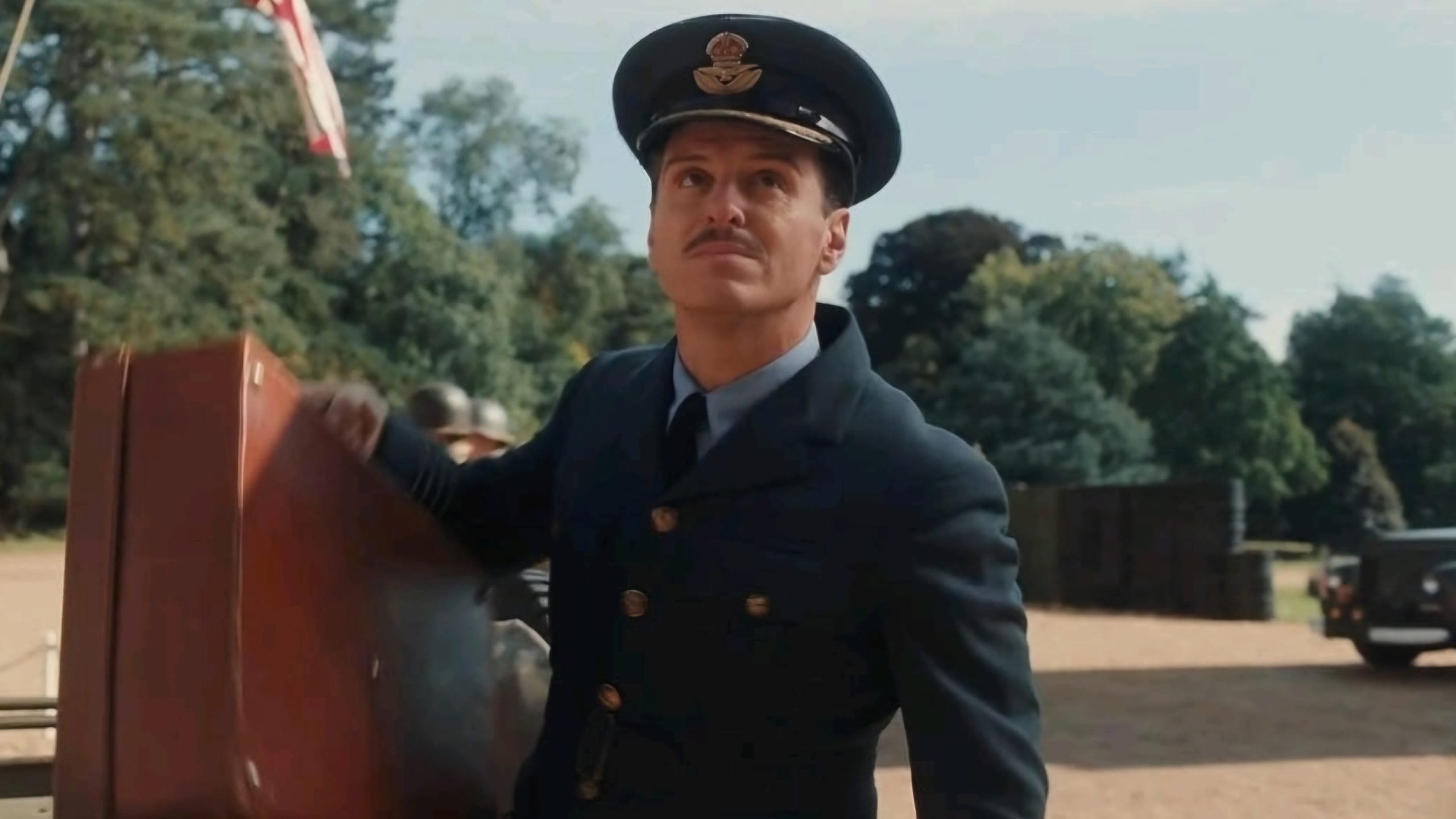 A man in a dark blue military uniform and cap stands outdoors, holding a large brown suitcase, with trees and equipment visible in the background.