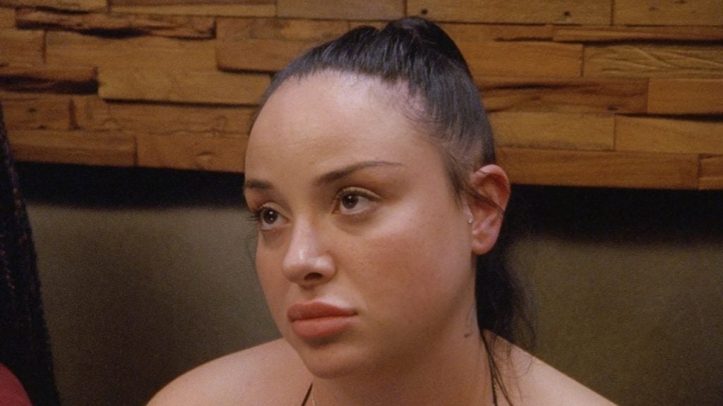 A person with long dark hair in a ponytail looks off-camera with a neutral expression, sitting indoors against a wooden wall.