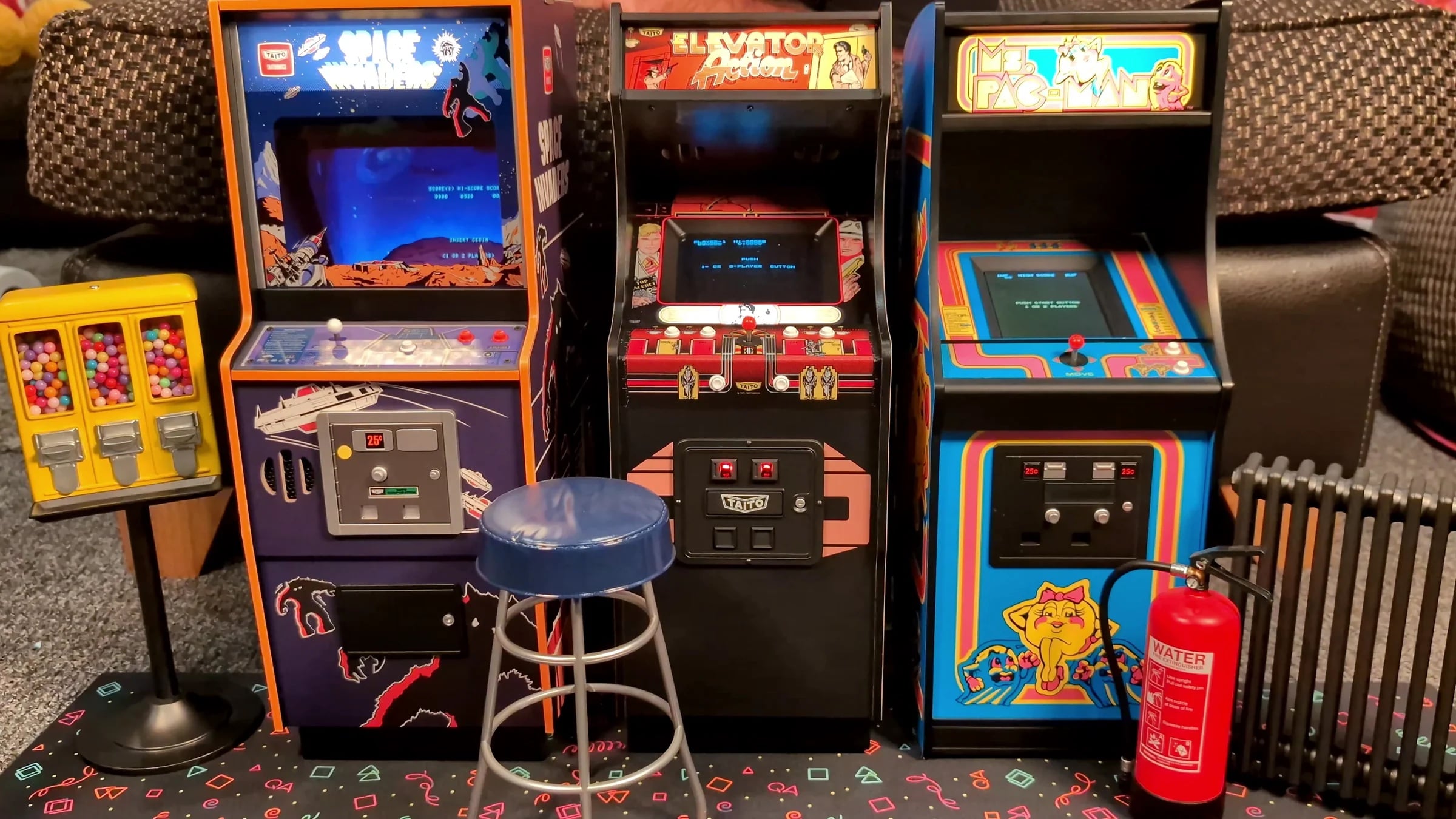 Three vintage arcade game machines—Ms. Pac-Man, Elevator Action, and Pac-Man—stand side by side, with a blue stool and a gumball machine in front of them on a patterned carpet.