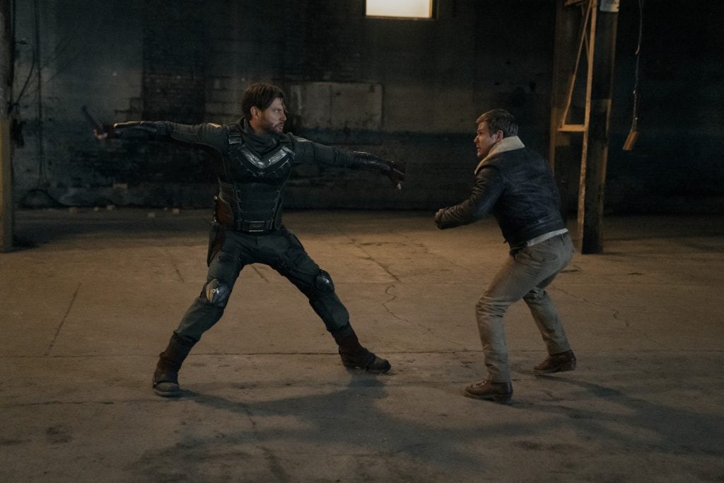 Two men face off in a dimly lit, empty warehouse; one wears tactical armor with arms spread, while the other stands ready, dressed in casual clothes and a jacket.