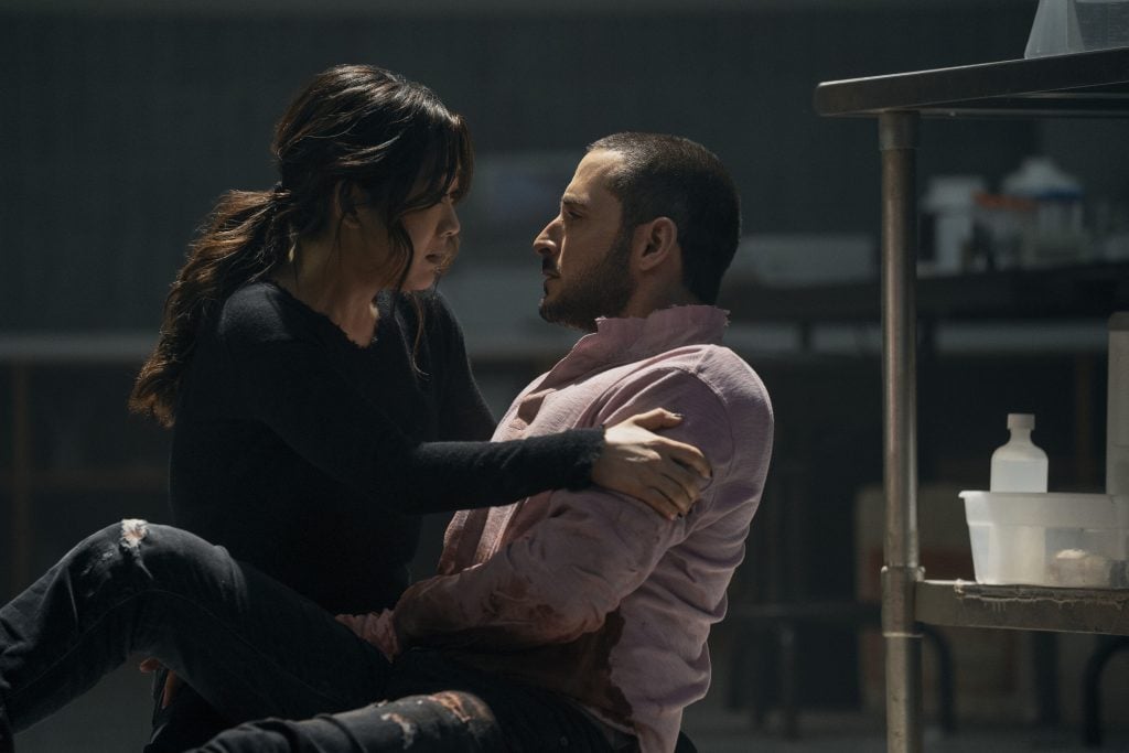 A woman holds an injured man in her arms on the floor of a dimly lit room, both looking at each other with serious expressions.