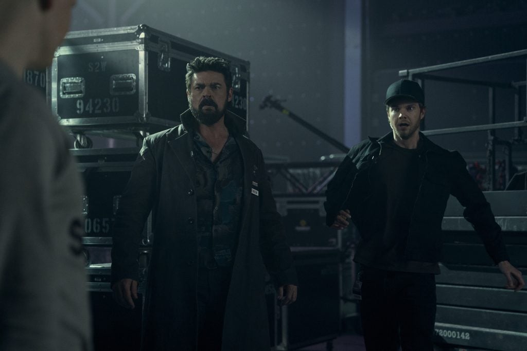 Two men stand in a dimly lit industrial setting with metal cases in the background; one looks serious, the other appears surprised or alarmed.