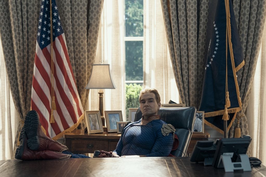 A man in a superhero costume sits at a large desk with feet up, in an office with American flags, family photos, and curtains framing a window behind him.