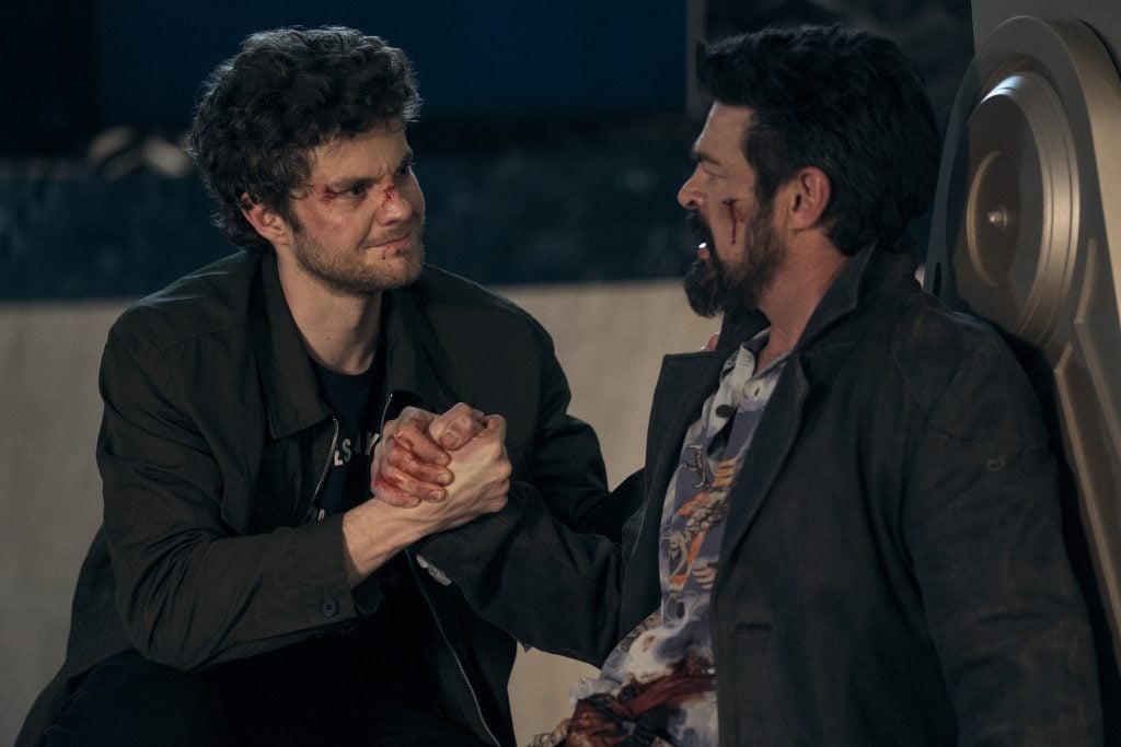 Two men with bloodied faces clasp hands and look at each other seriously, sitting on the ground in a dimly lit setting.