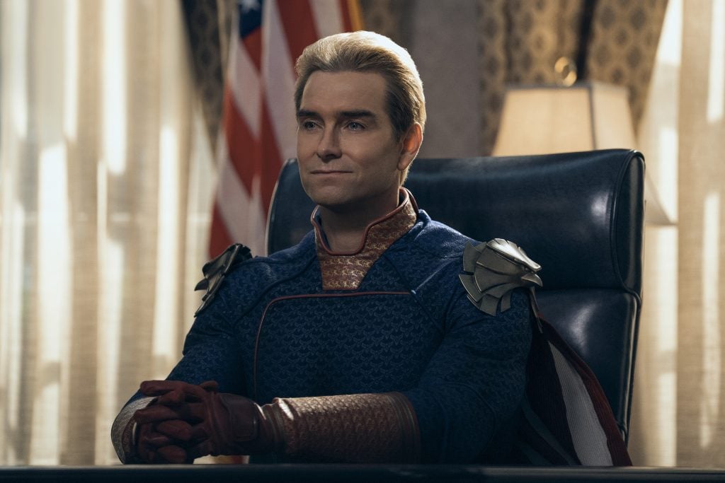 A man in a superhero costume sits at a desk with his hands clasped, an American flag and lamp in the background.