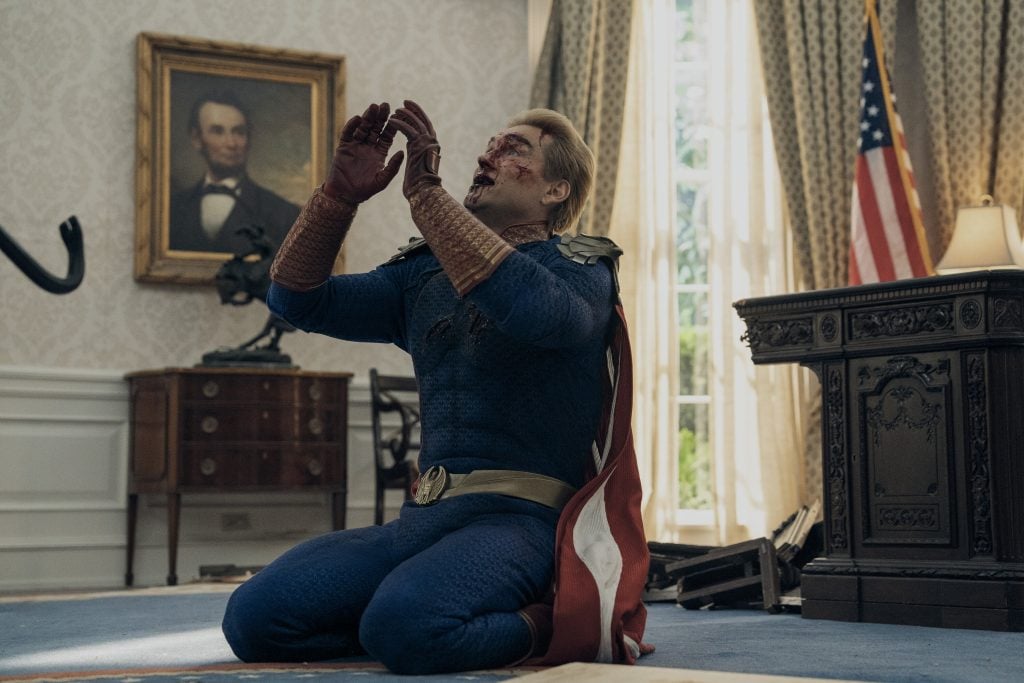 A man in a superhero costume kneels on the floor of an office resembling the Oval Office, with a bloody face and hands raised. An American flag and Abraham Lincoln portrait are visible.