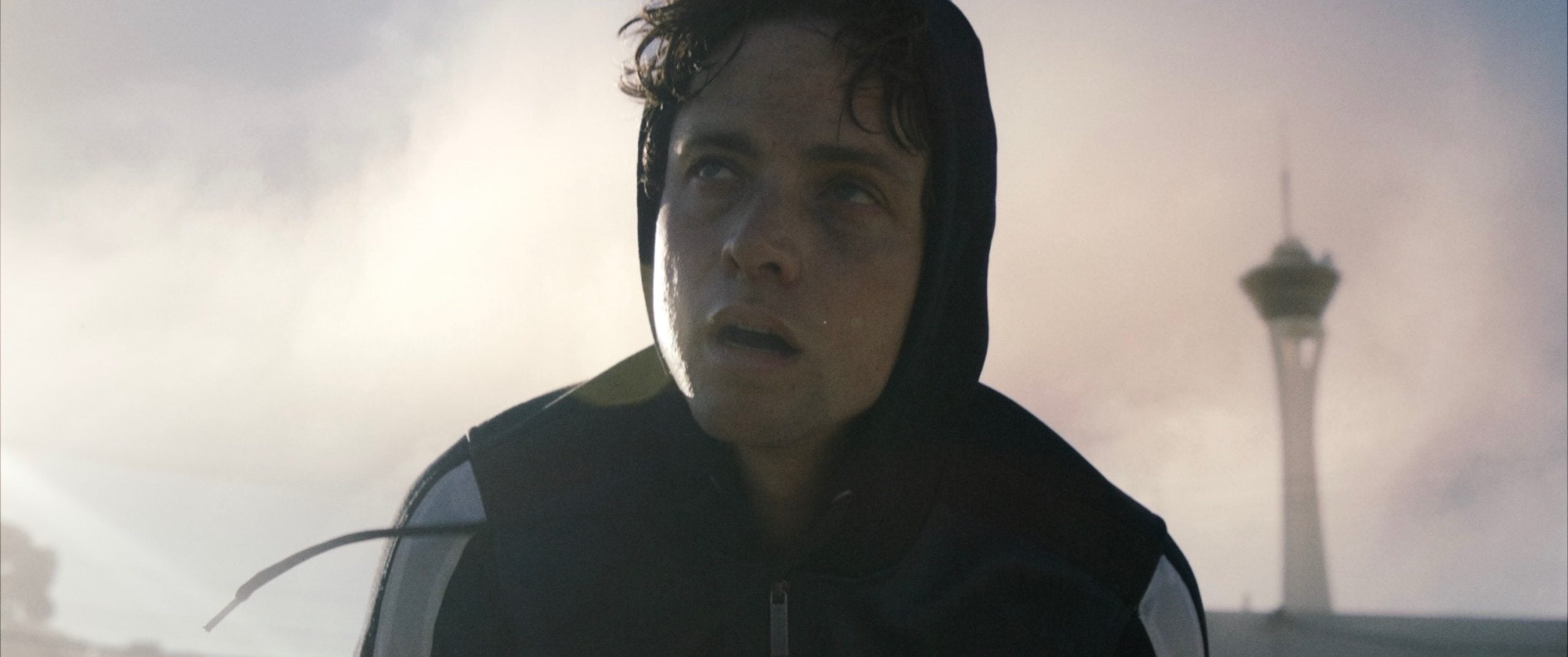 A person wearing a hooded jacket looks upward with a tired expression outdoors, with a tall tower and a cloudy sky in the background.