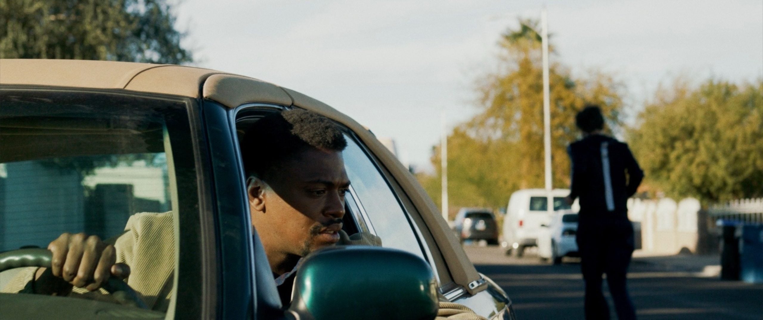 A man sits in a parked car looking out the window while another person walks away down a suburban street.