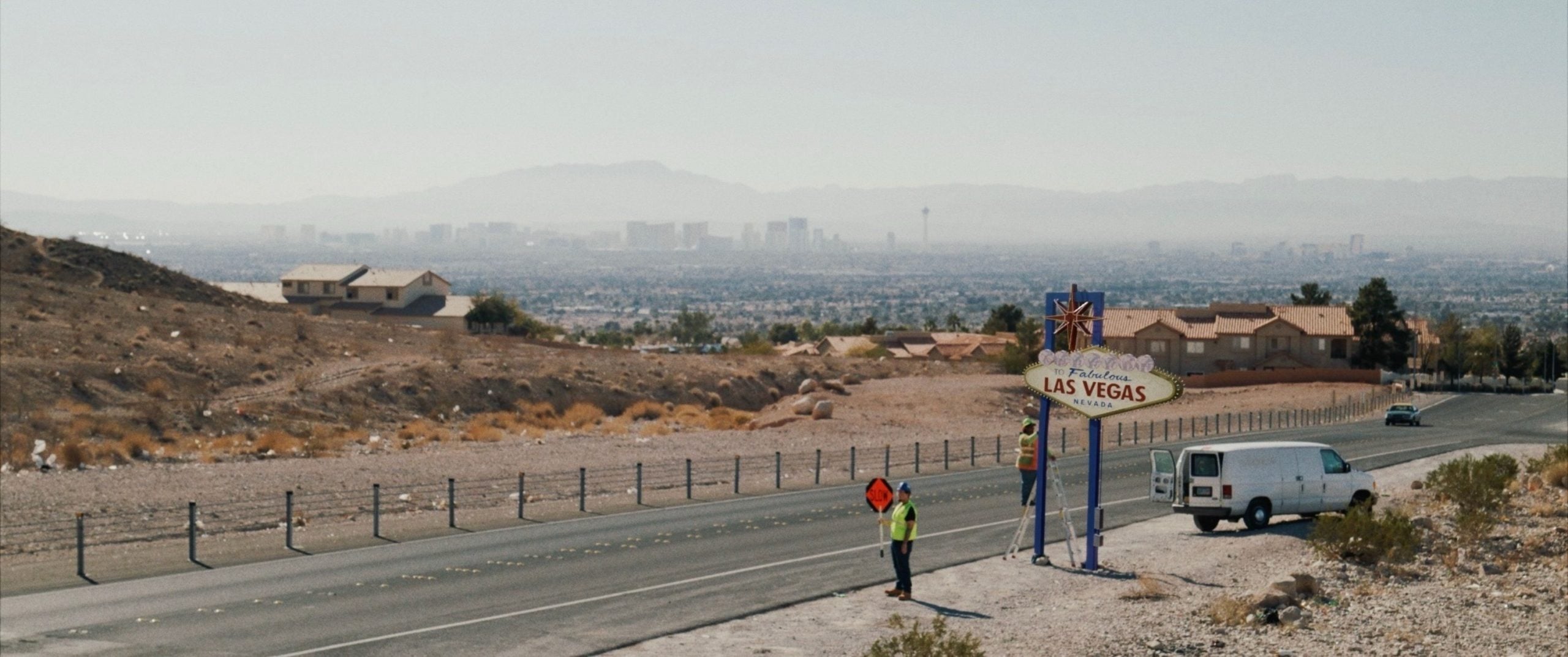 A person in a safety vest stands beside a "Welcome to Las Vegas" sign on a roadside with a parked white van; city skyline visible in the hazy distance.