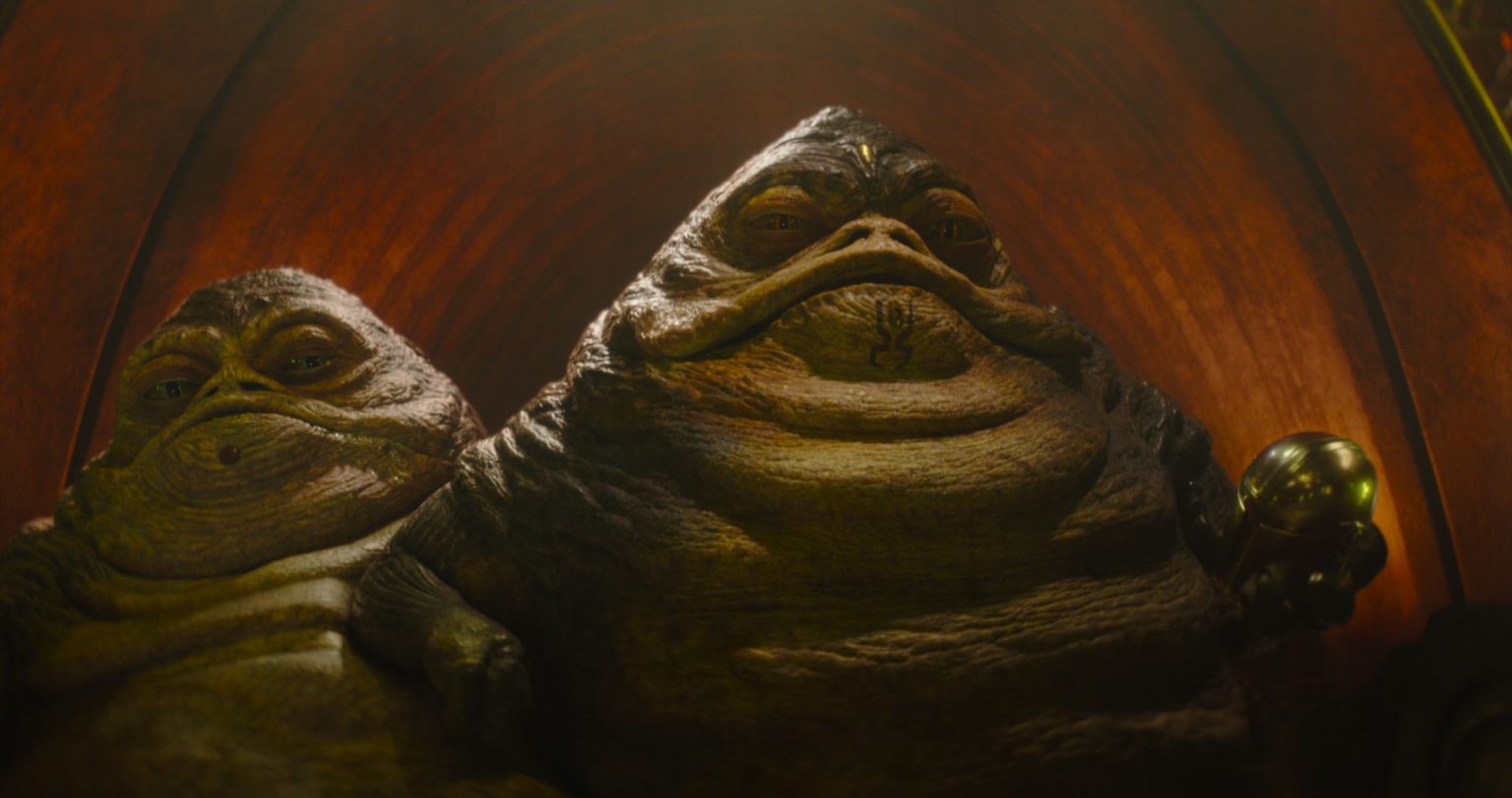 Two large, slug-like alien creatures with wrinkled skin and serious expressions sit side by side in a dimly lit, arched space.
