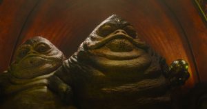 Two large, slug-like alien creatures with wrinkled skin and serious expressions sit side by side in a dimly lit, arched space.