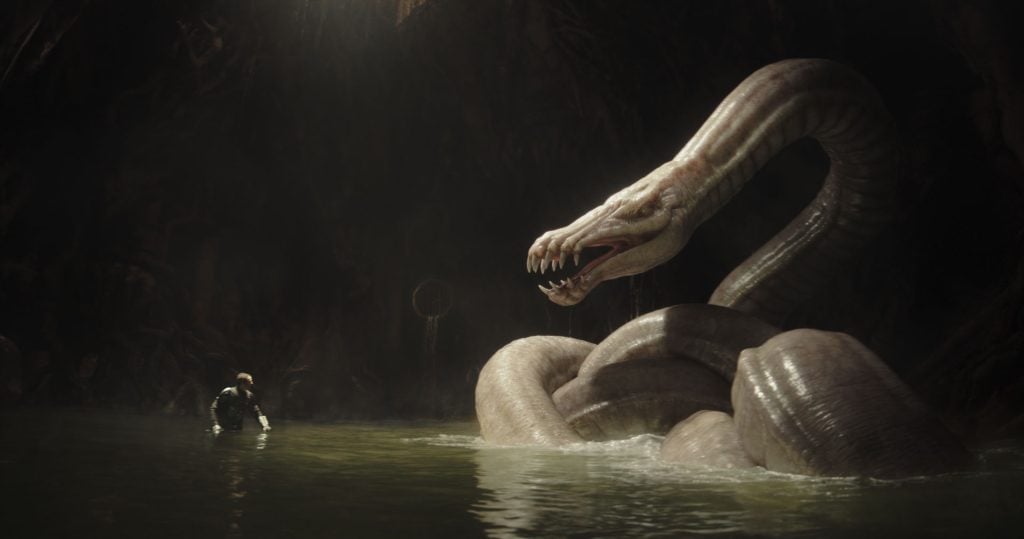 A person stands in shallow water facing a giant, menacing serpent with its mouth open and fangs exposed in a dark, cavernous setting.