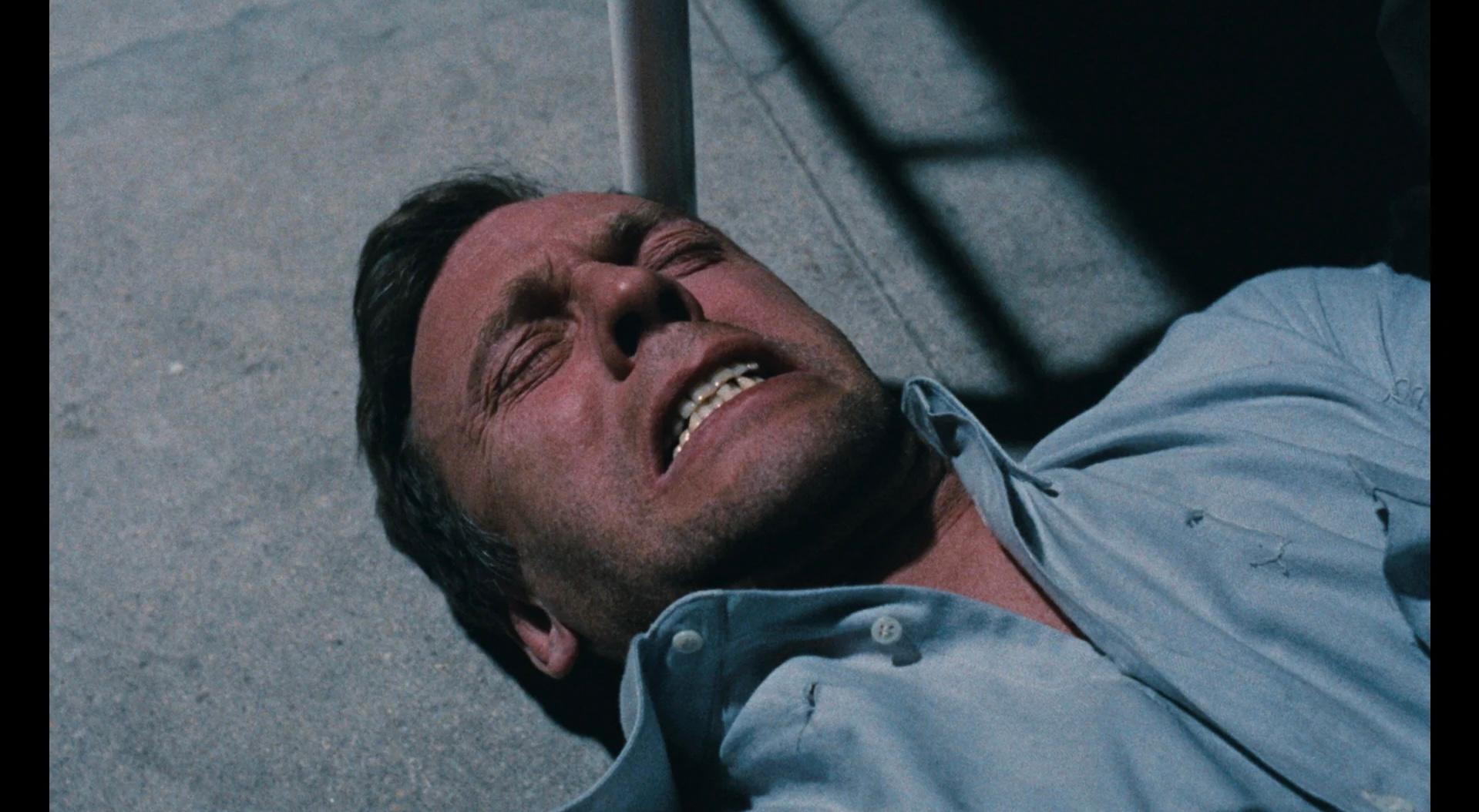 A man in a light blue shirt lies on the ground, grimacing in pain or distress under harsh lighting.