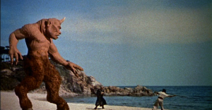 A large, horned creature stands on a beach as several people point or react to its presence, with the ocean and rocky cliffs in the background.