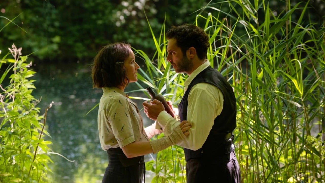 A woman with a bandaged arm stands facing a man holding a knife in a lush, green outdoor setting near water.