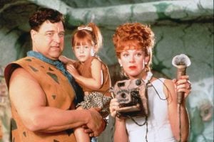 A man in a stone-age outfit holds a young girl, while a woman with red hair holds a prehistoric-style phone and microphone in a cave-like setting.