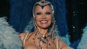 A performer wearing an ornate headdress, jeweled necklace, and blue feathered costume smiles on stage.