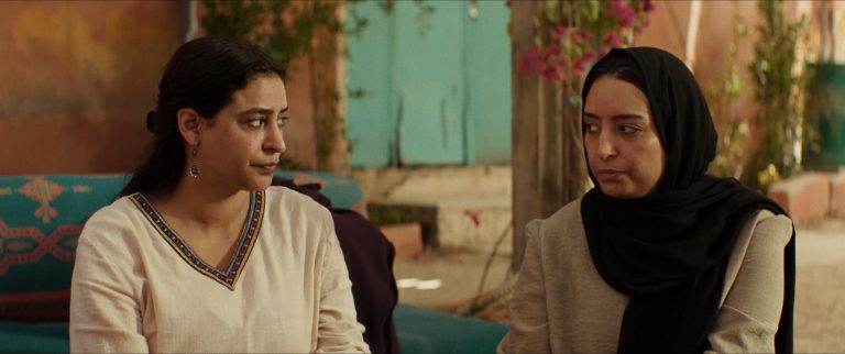 Two women sit side by side outdoors, facing each other with serious expressions. One wears a light blouse, the other a beige top with a black headscarf. Plants and a blue door are in the background.