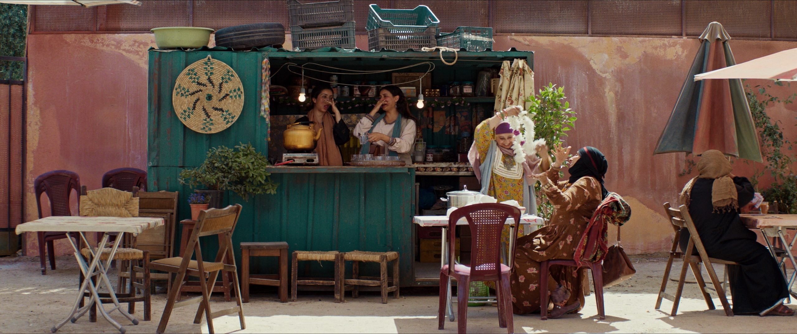 A small outdoor café with people sitting at tables and two women working inside a green food stall, set against a peach-colored wall.