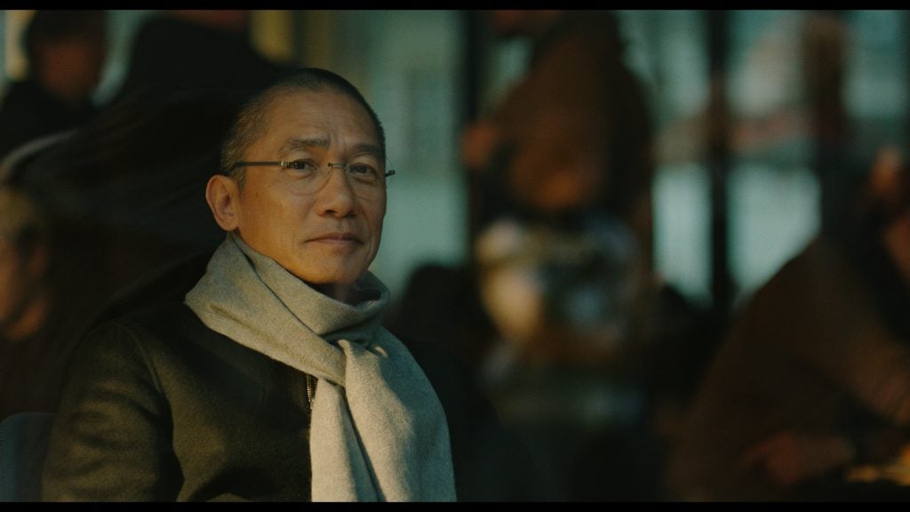 An older man wearing glasses, a scarf, and a dark coat sits indoors with a calm expression, while other people are blurred in the background.