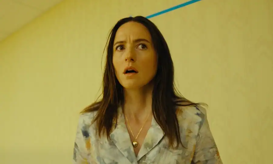 Woman with long brown hair wearing a patterned shirt and necklace stands in a yellow hallway, looking ahead with a concerned expression.